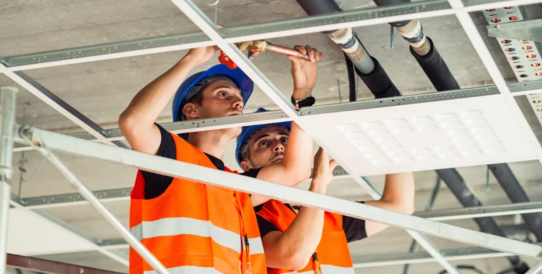 Two construction workers in safety vests and hard hats are installing pipes in a ceiling framework.